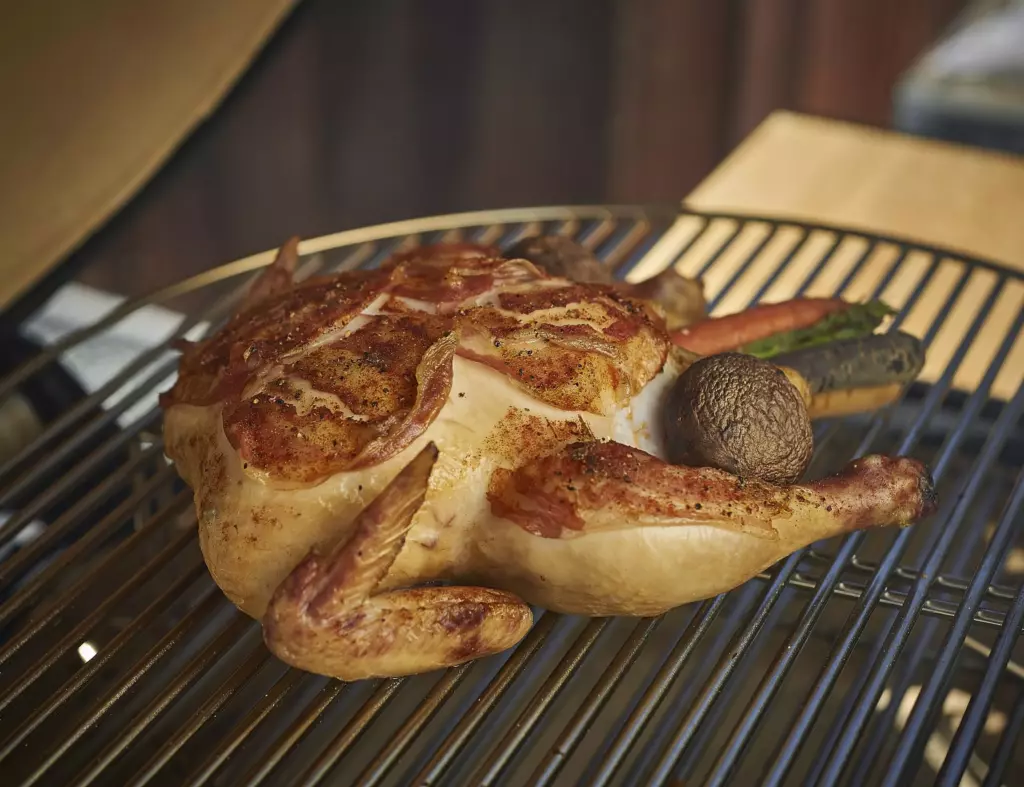 A fully cooked, small, bacon-wrapped whole chicken sits on a grill cooking surface with a mushroom and carrots surrounding it.