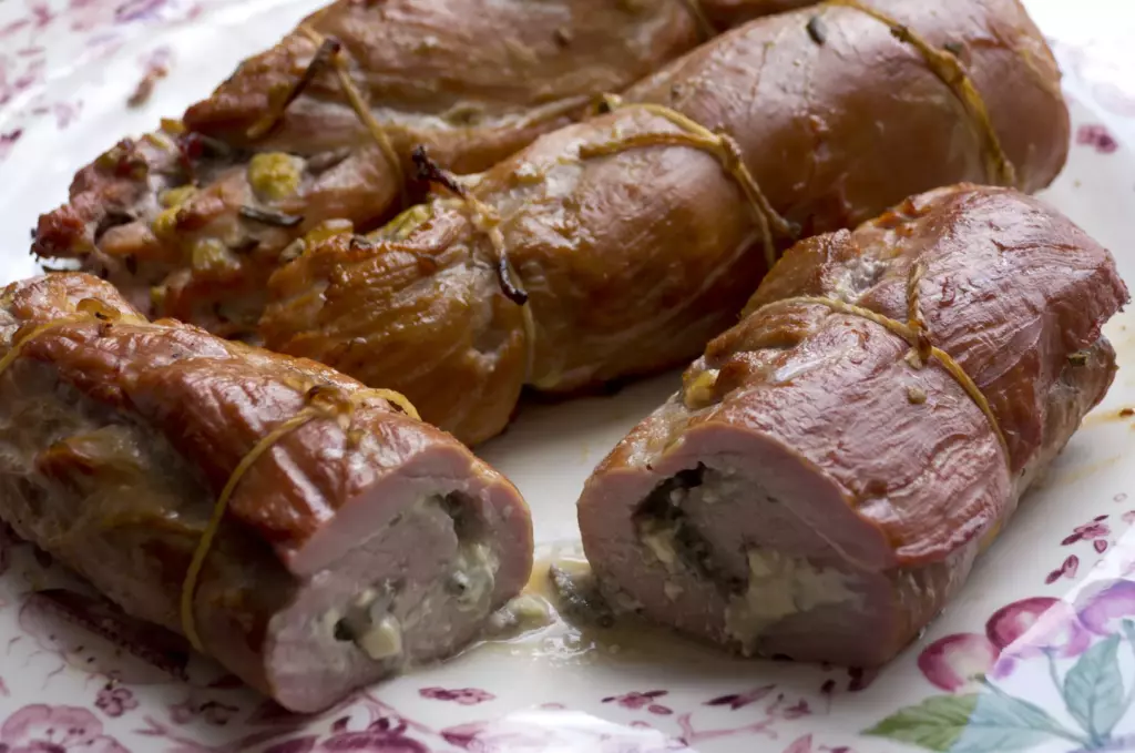 Four stuffed tenderloins are shown on a plate, still tied up with waxed twine. The stuffing is coming out of two of the pieces.