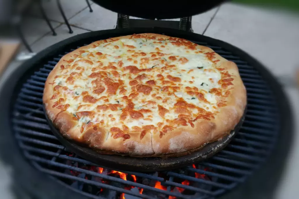 A cheese and veggie pizza is seen cooking inside of a kamado grill. The cheese is golden brown, and the charcoal underneath the pizza stone is red hot. It looks ready to eat.